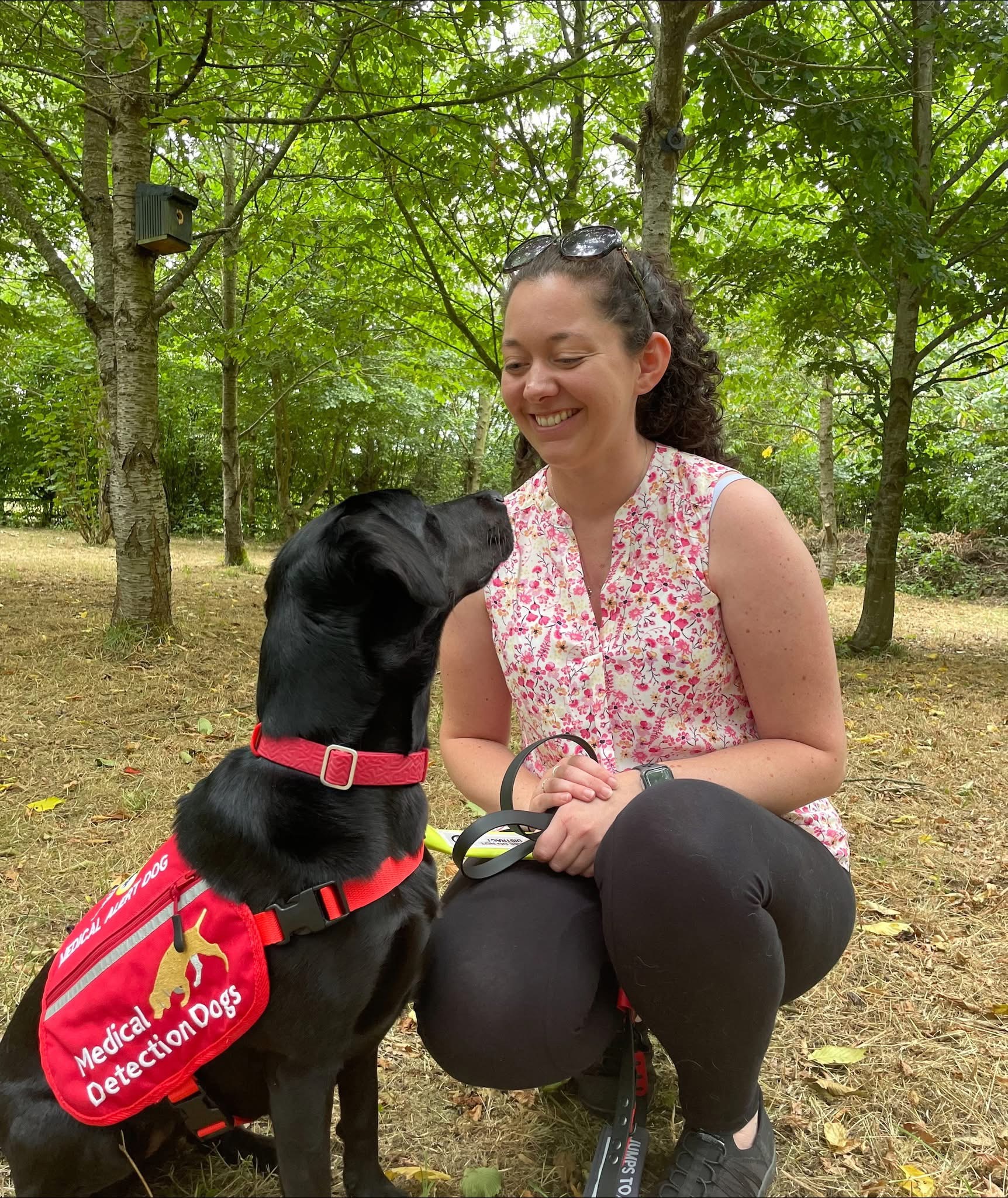 Laura, wearing a floral sleeveless top, kneels in a leafy outdoor setting, smiling at her black Labrador, Mae, who is wearing a red ‘Medical Detection Dog’ vest.