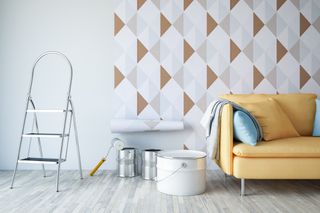 A white room with brown, blue, white and grey chevron patterned wallpaper being glued to the wall, along with tins, a stepladder and a yellow sofa.