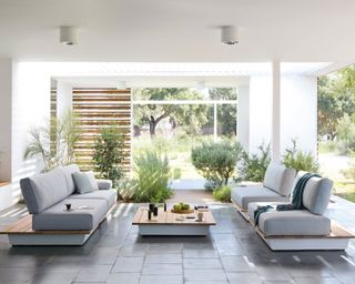 Indoor-outdoor room with two gray, white and wood sofas, tiled flooring, planting, wooden paneling in background, white painted walls