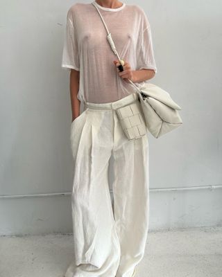 A model wearing a sheer white T-shirt with white linen pants and two white bags.