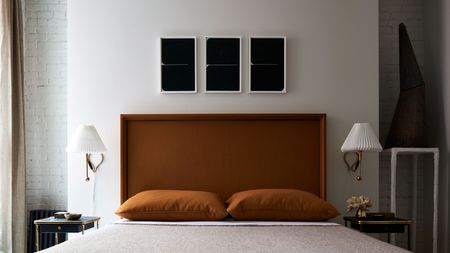 minimal white bedroom with orange headboard accent and bed linen with lamp sconces and three artwork hanging above bed