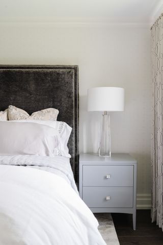 white bedroom with black velvet headboard, very pale gray bedding and side table, white and glass table lamp, rug, dark wood floor