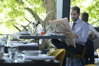 Jesse Bloom (Jay Duplass) sits at a window seat in a hotel restaurant, wearing a shirt and tie and reading the Financial Times