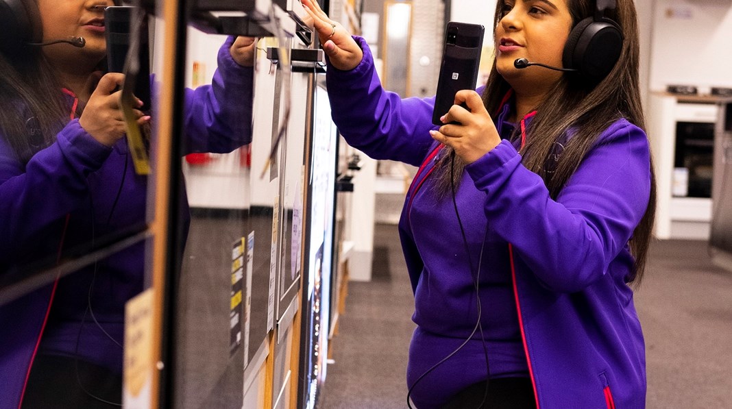 A Currys store employee wearing a purple zip-up hoodie and a headset holds a smartphone while reaching to adjust a shelf or product display.