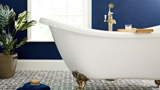 White rolltop freestanding bath with ornate brass feet set against a dark blue wall and decorative Victorian tiles and plant