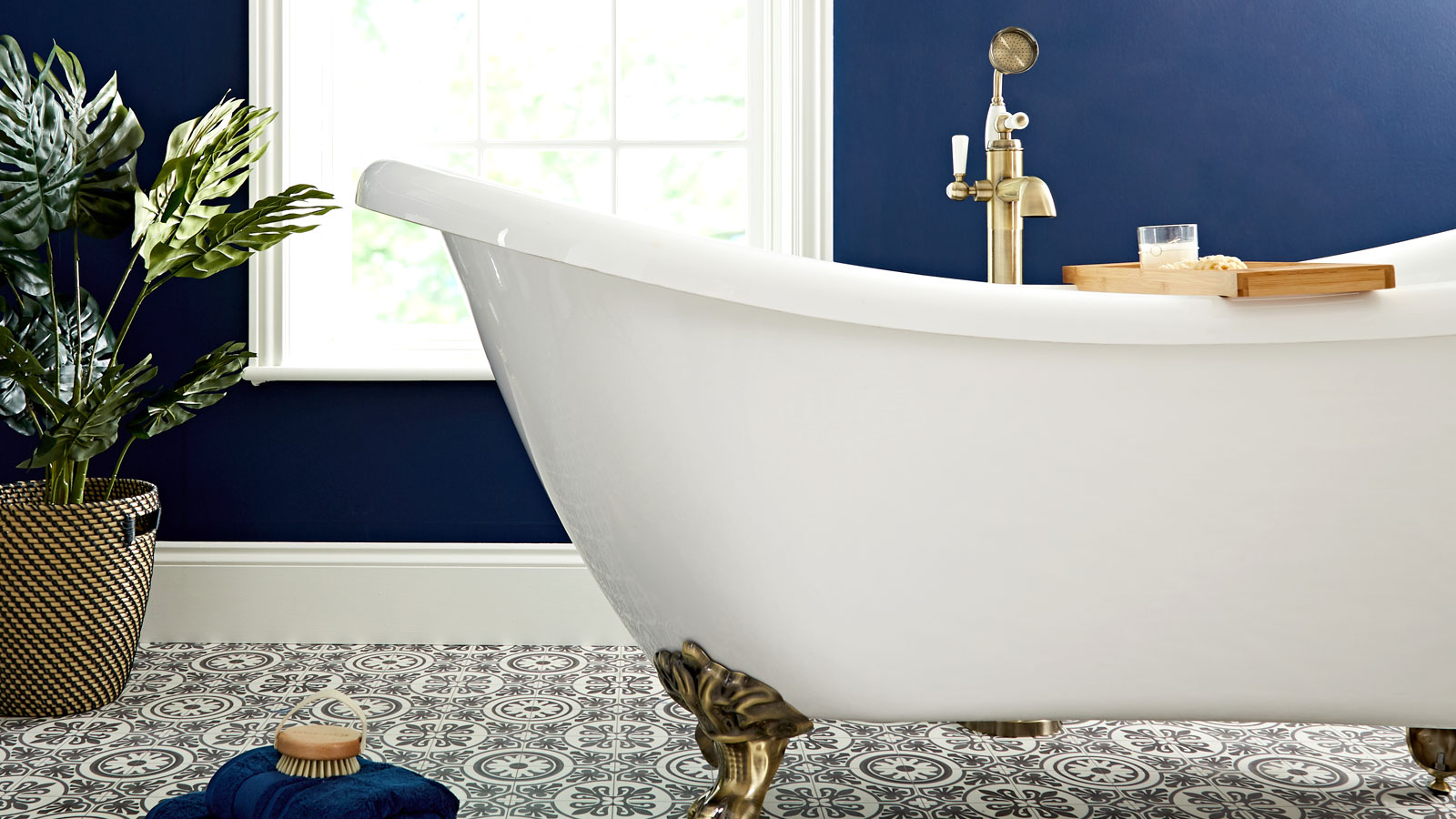 White rolltop freestanding bath with ornate brass feet set against a dark blue wall and decorative Victorian tiles and plant