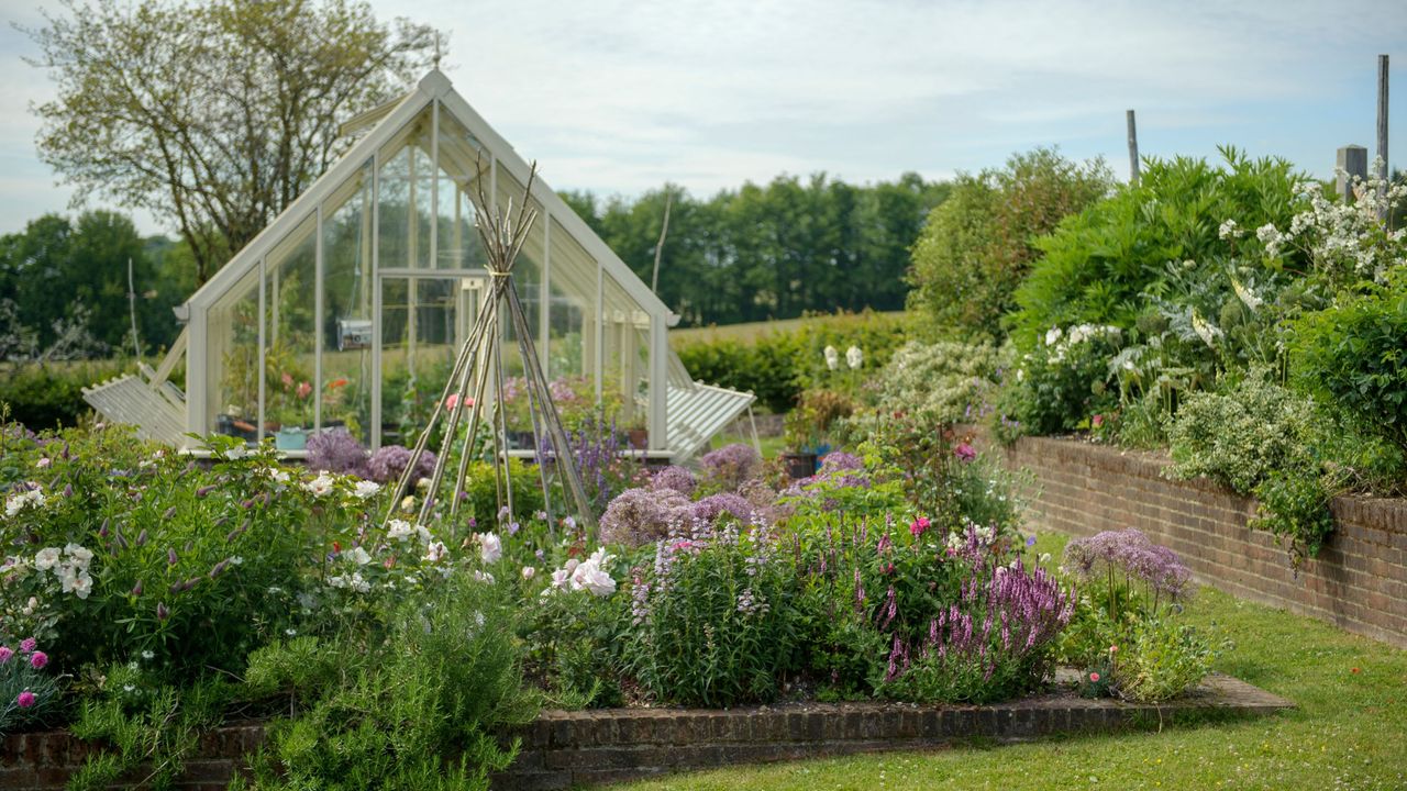 A lush garden filled with flower beds and raised borders, with a large white greenhouse in the background