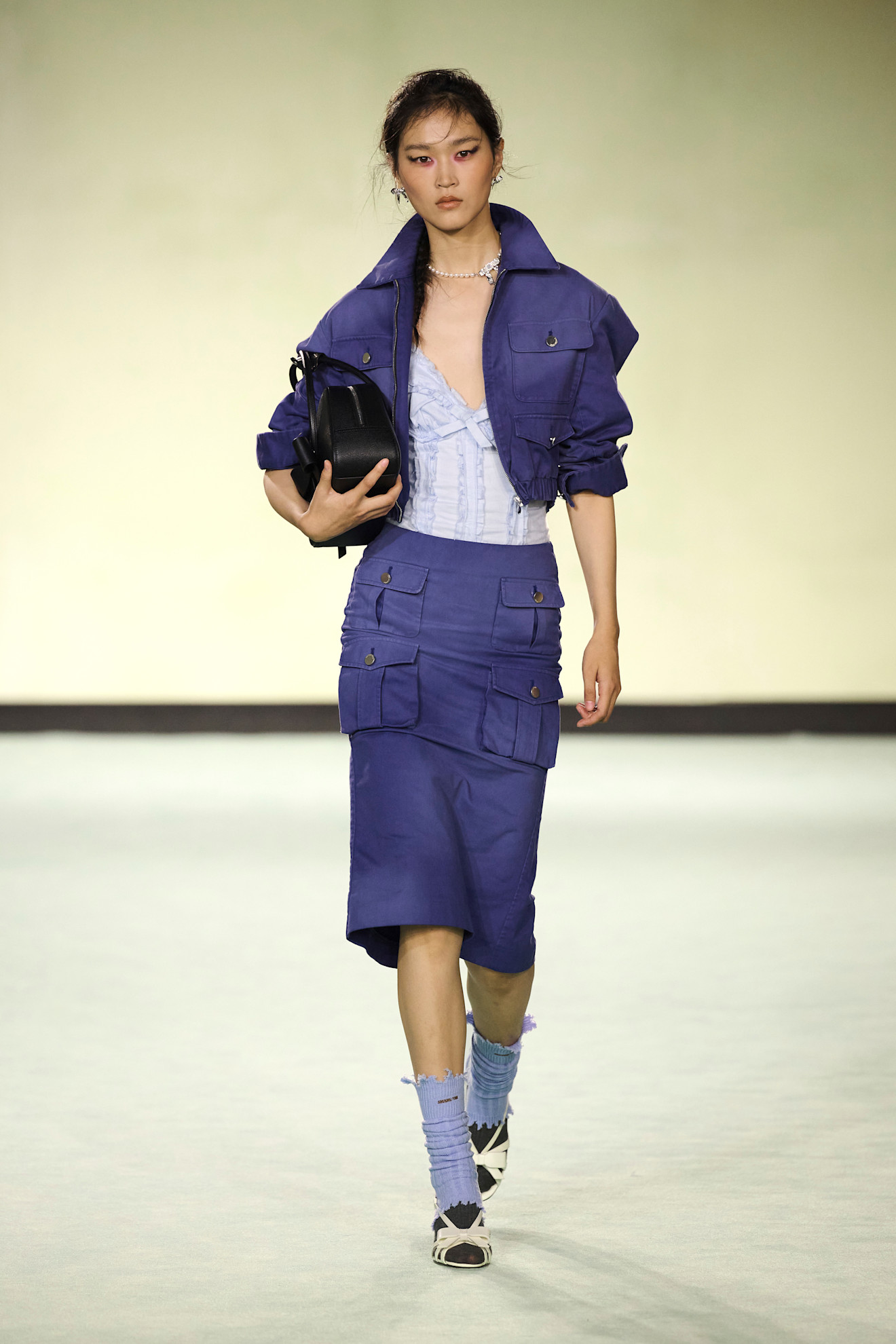 A Shushu/Tong model wearing a purple cargo jacket layered over a light lilac lace bodysuit, a dark purple cargo skirt, white sandals, brown socks, lilac leg warmers, a black bowler bag, and a pearl necklace.