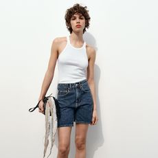 Model wears whit tank top and longer deep blue denim shorts while carrying a handbag