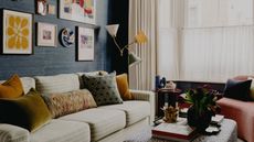 A cozy living room with dark blue walls with a gallery wall, a bay window with cafe curtains and cream curtains, a pale green striped sofa with colorful cushions and a leopard print ottoman.