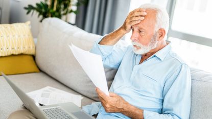 An older man holds a hand to his head in shock as he looks at paperwork while sitting on his sofa.