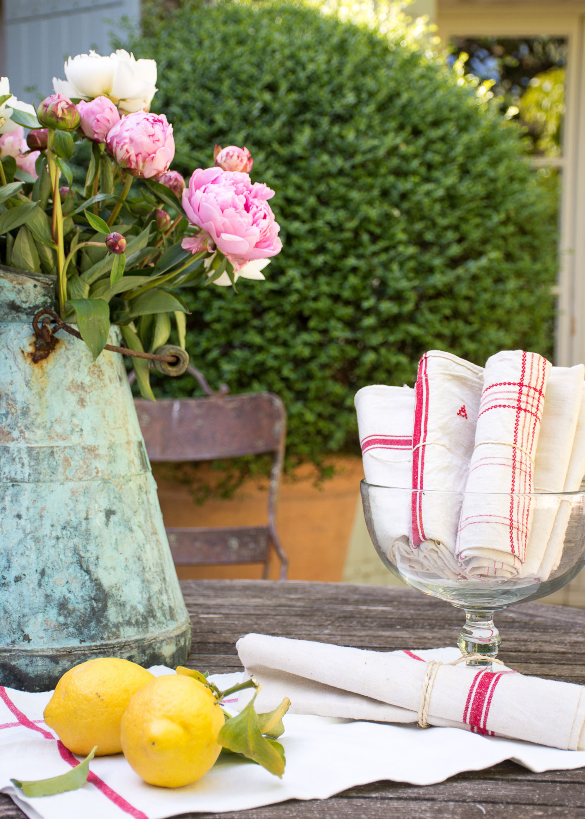 An antique watering can with peonies on a garden table with a glass pedestal bowl