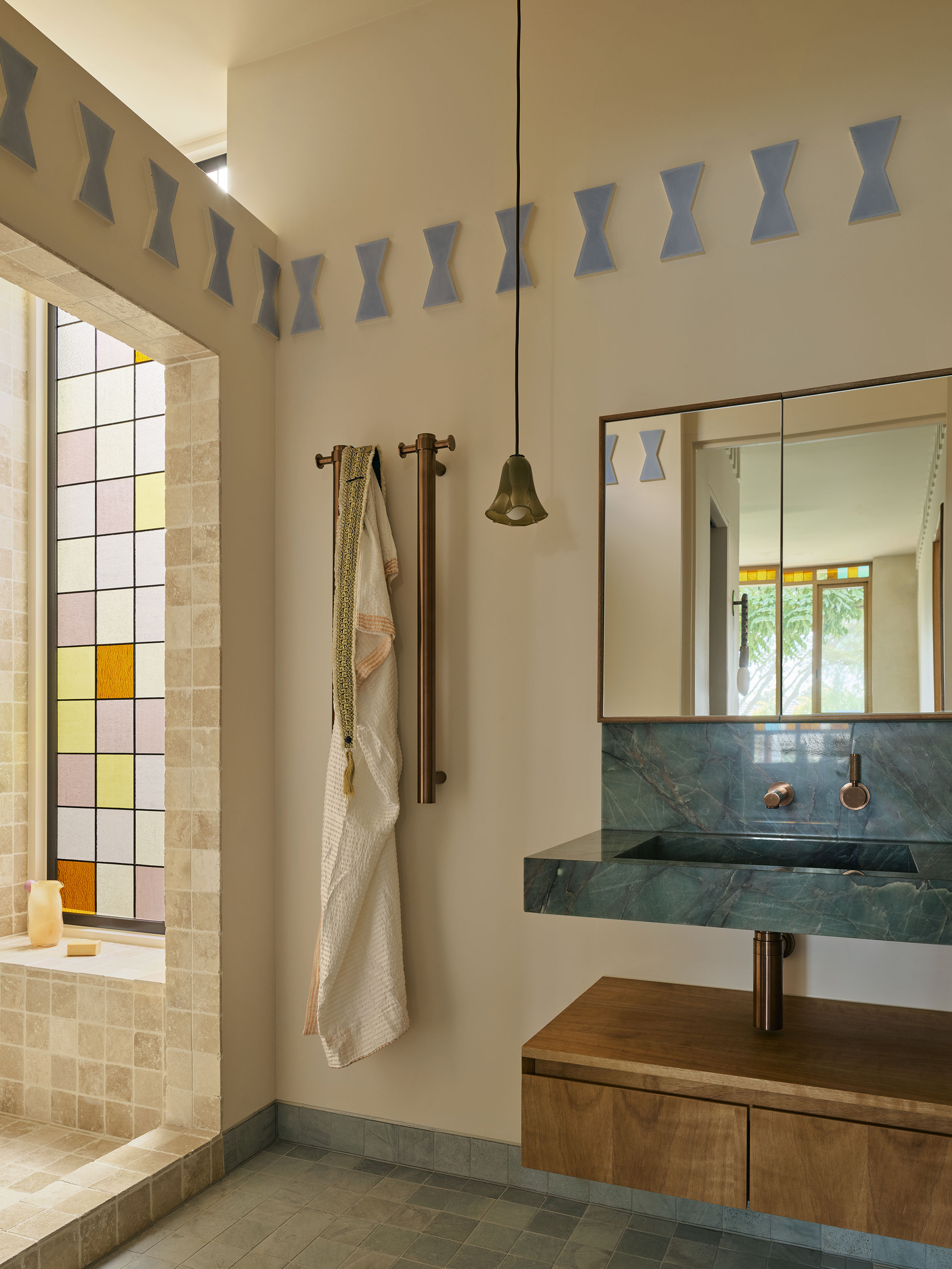 modern bathroom with cream walls, green marble floating sink with timber cabinets, decorative wall molding, tiled shower enclosure with colorful stained glass window and two vertical towel holders with robe hanging