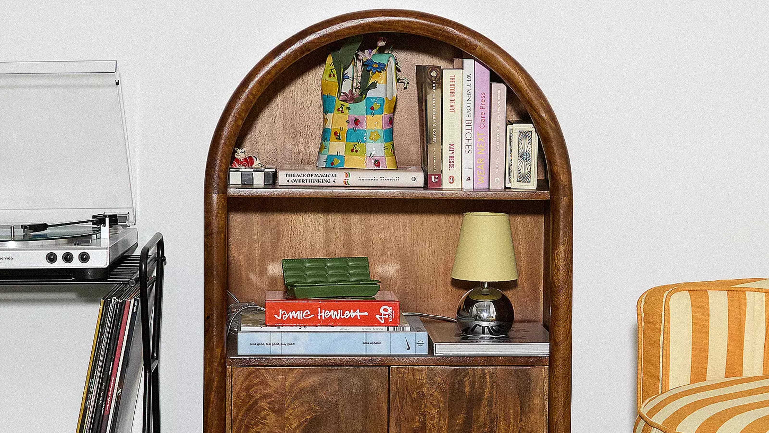 A living room with an Urban Outfitters arched wooden display cabinet filled with books and a chrome table lamp