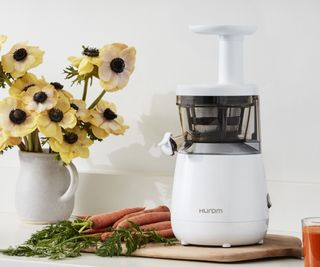 Hurom slow juicer on a chopping board with carrots and a vase of flowers in he background
