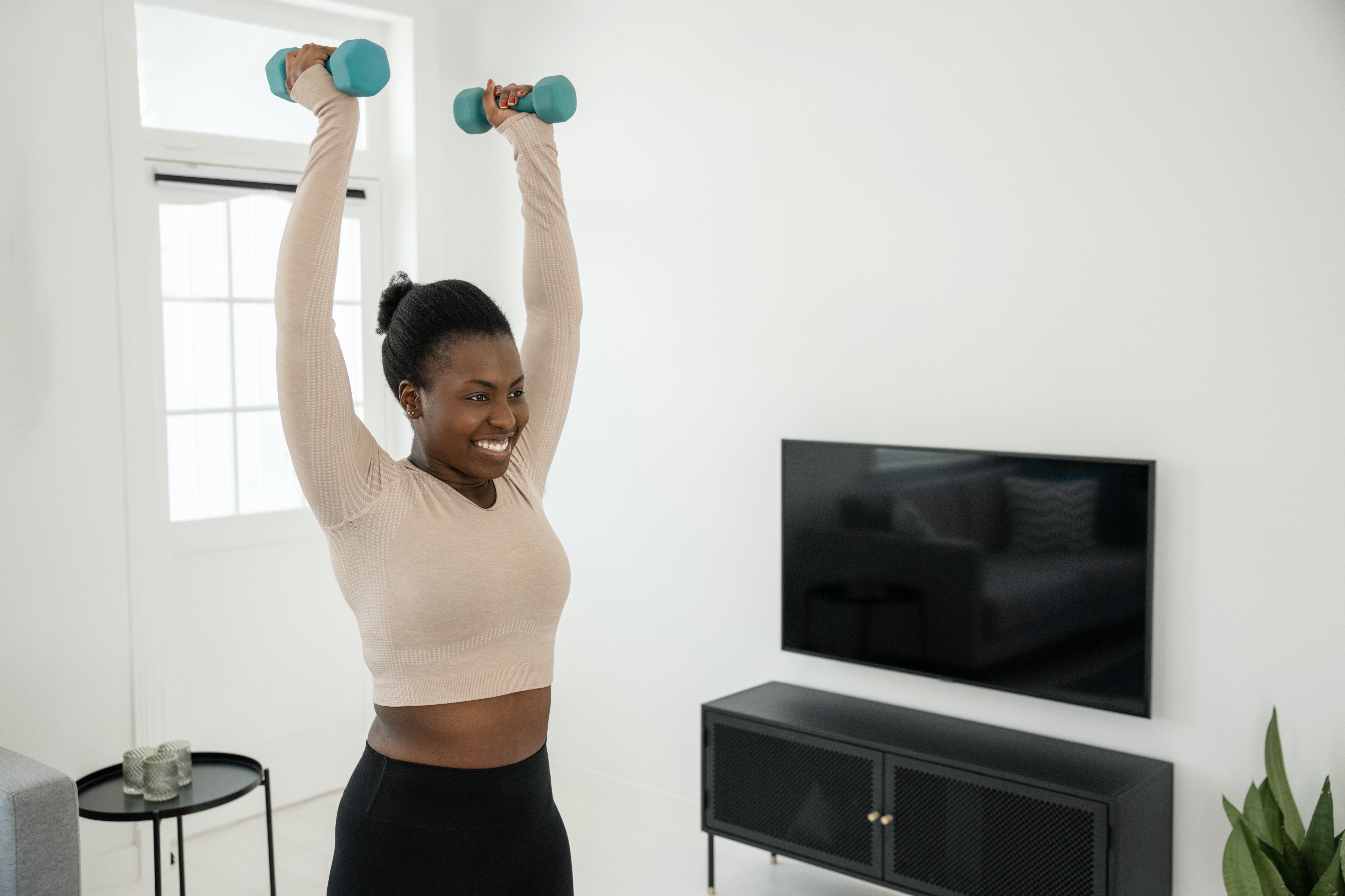 A smiling woman in long-sleeved crop top and leggings stands holding a pair of dumbbells overhead in her living room. Behind her we see a TV, plant and table.
