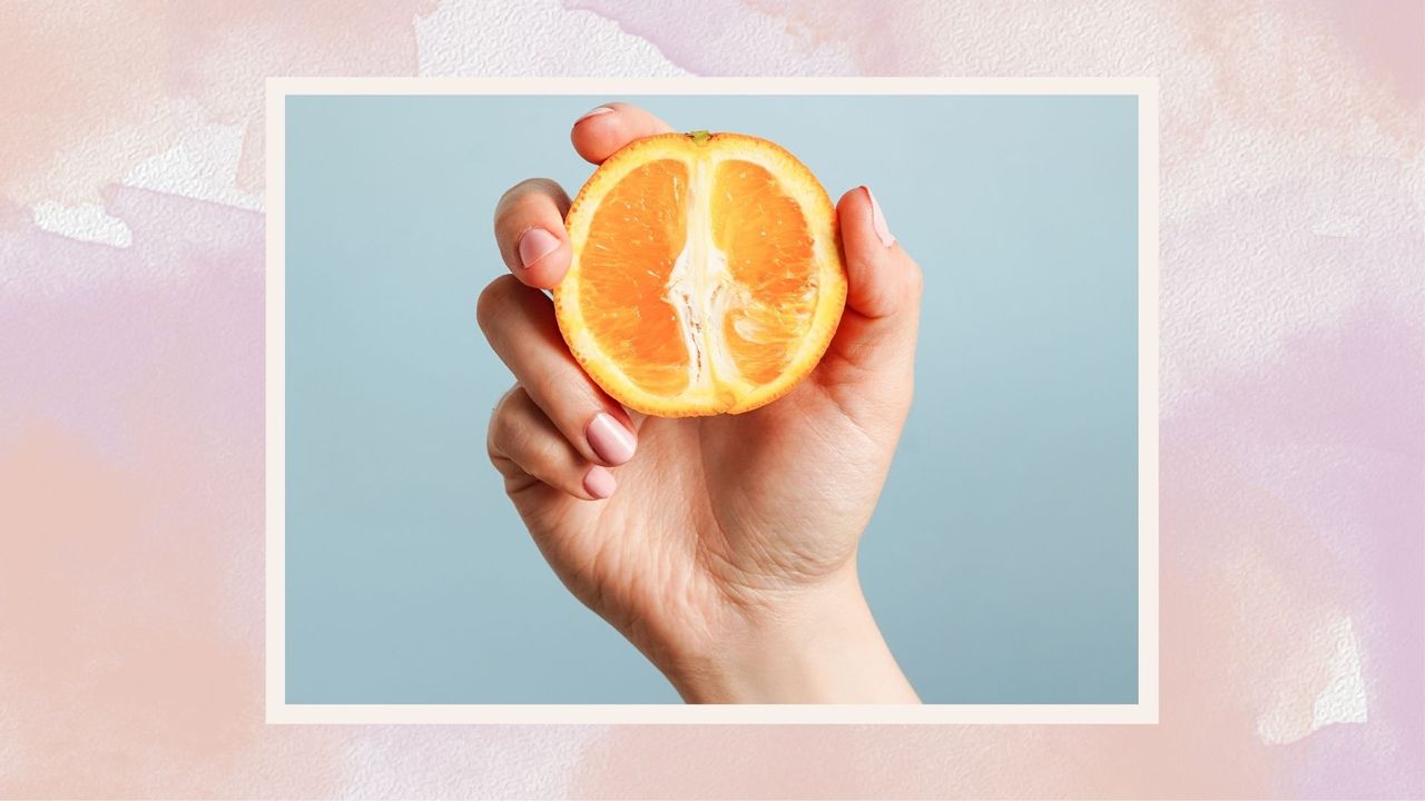 A woman&#039;s hand holding an orange on a pink watercolour backdrop