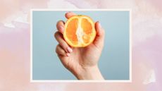 A woman's hand holding an orange on a pink watercolour backdrop