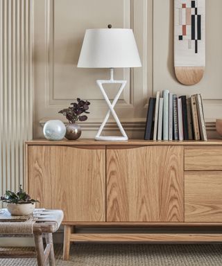 An off white panelled wall, wood finish sideboard with geometric white lamp, round metal vase with dried purple leaves, white glass ball, and modern art on the wall.
