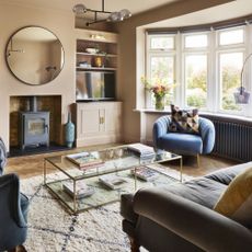 Cosy warm neutral living room with bay window, alcove storage, log burner, circle mirror, glass coffee table, textured rug and accent chair next to radiator