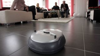 A robot vacuum cleaner cleans a tiled floor while people sit on couches in the background.