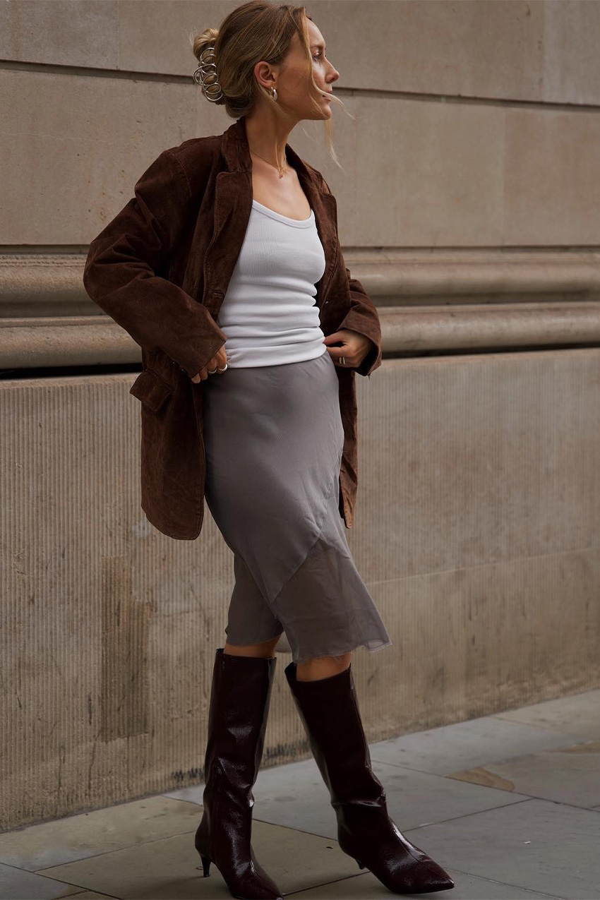 How to wear a slip skirt is shown in a photo of a woman&#039;s slip skirt outfit where she&#039;s standing outside on the sidewalk wearing a brown suede blazer over a white tank top with a gray slip skirt and brown knee-high boots