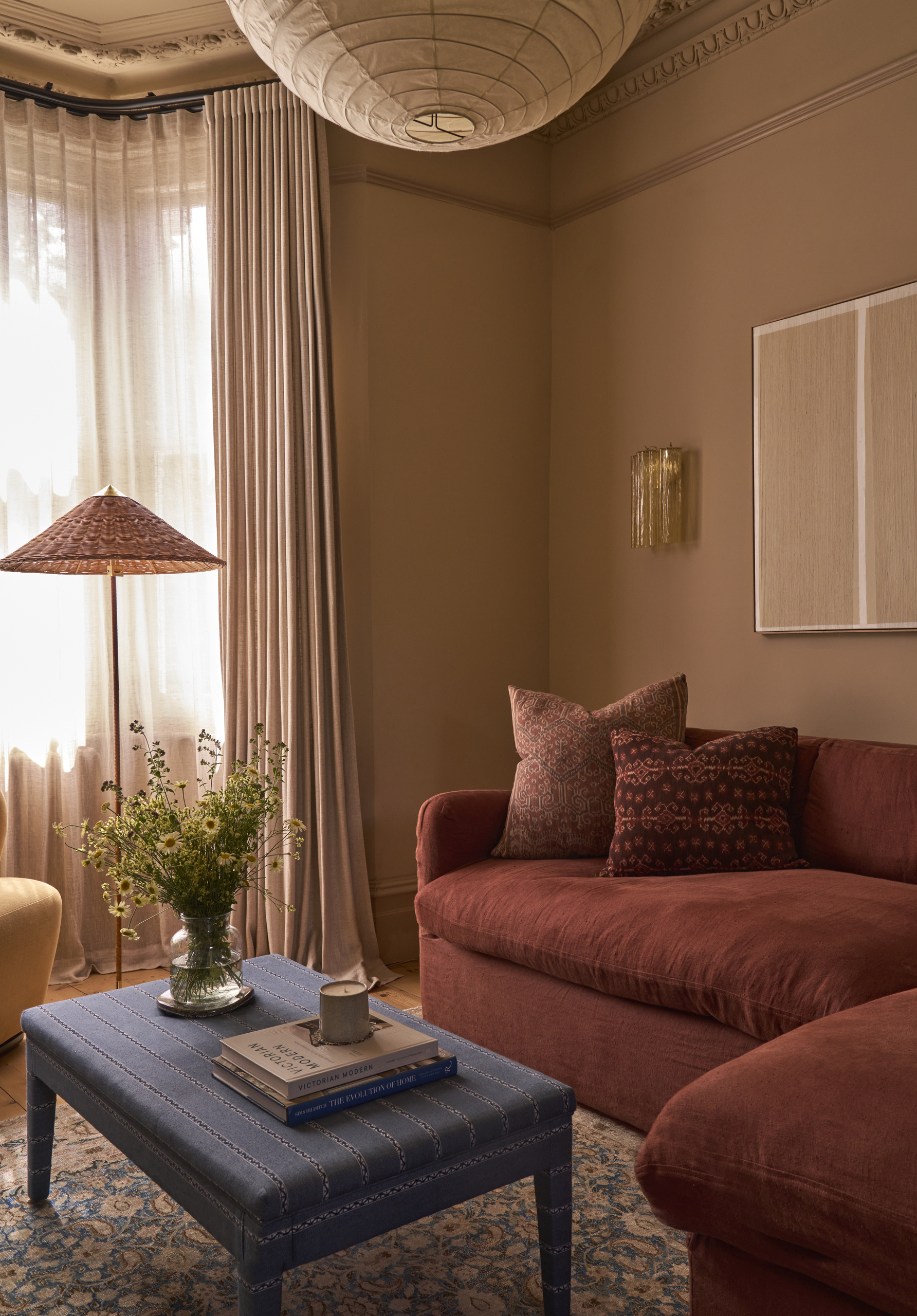 Image of a warm taupe color-drenched living room with molding around the ceiling, a red sofa, a blue upholstered coffee table, and a paper lantern.