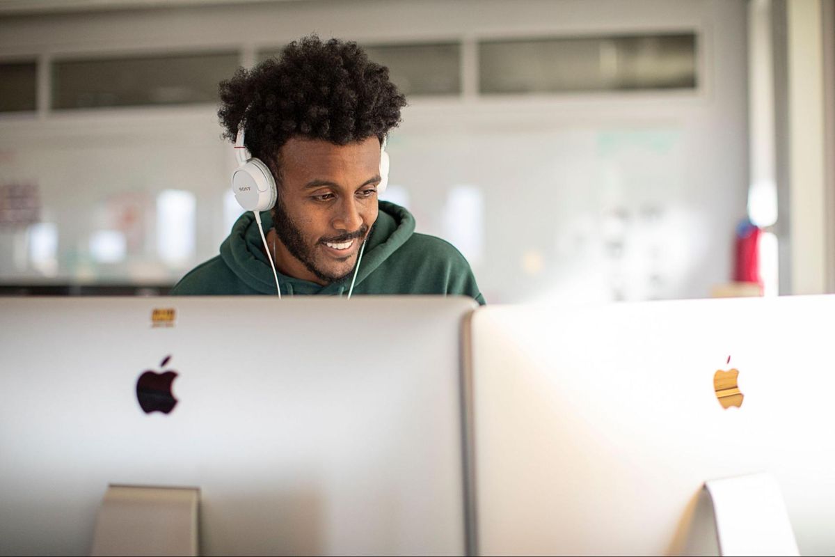 Black young adult with headphones sitting in front of two Apple monitors