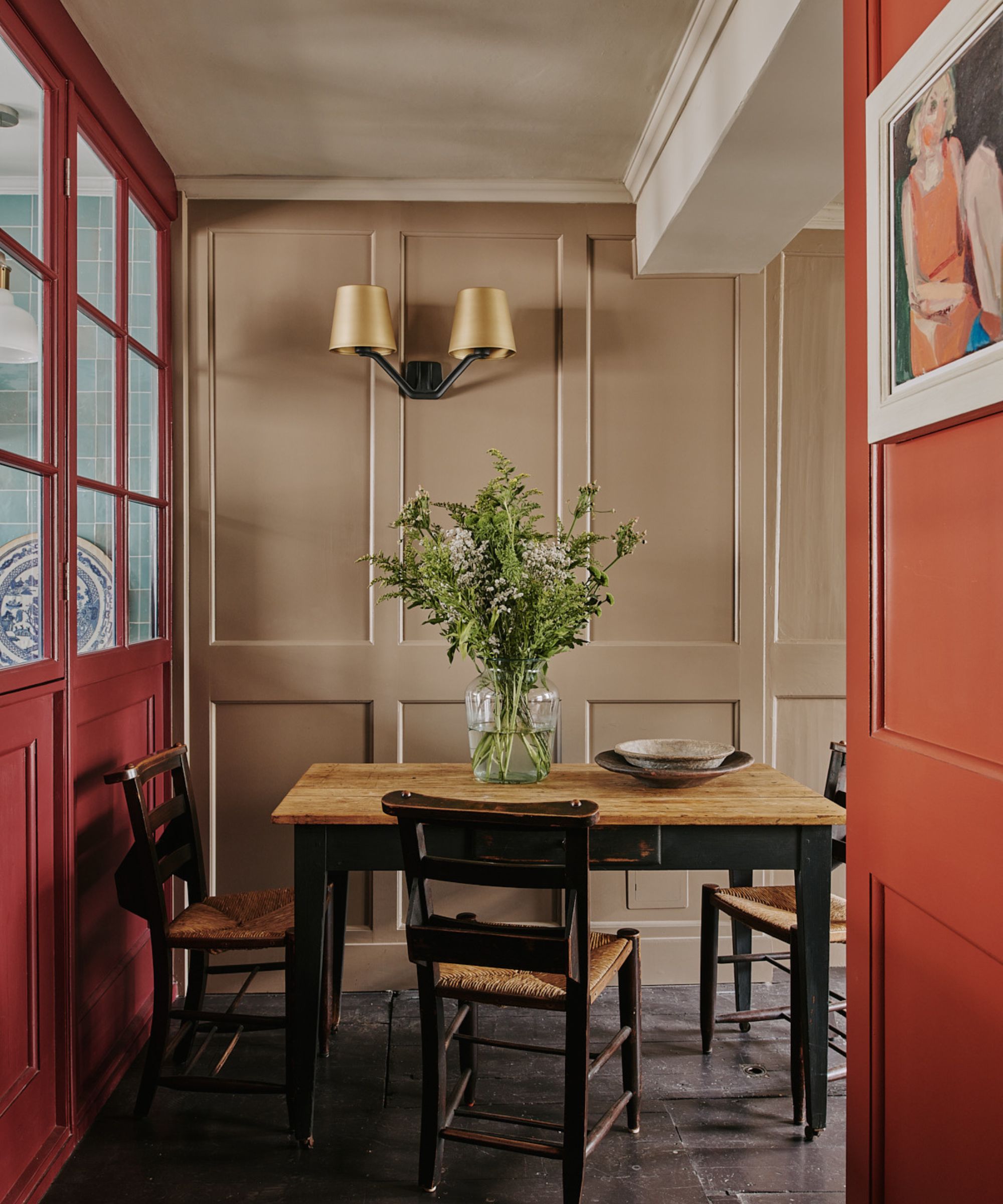 Dining room painted in Farrow &amp;amp; Ball's Mouse's Back, red glass doors, an antique wooden farmhouse table and wicker dining chairs, a large vase of foliage, and a double wall sconce.