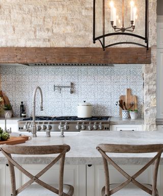 A French kitchen with a gray kitchen countertop with two dark wooden chairs underneath it with, with a silver oven with a white pot and a beige stone wall in front of it