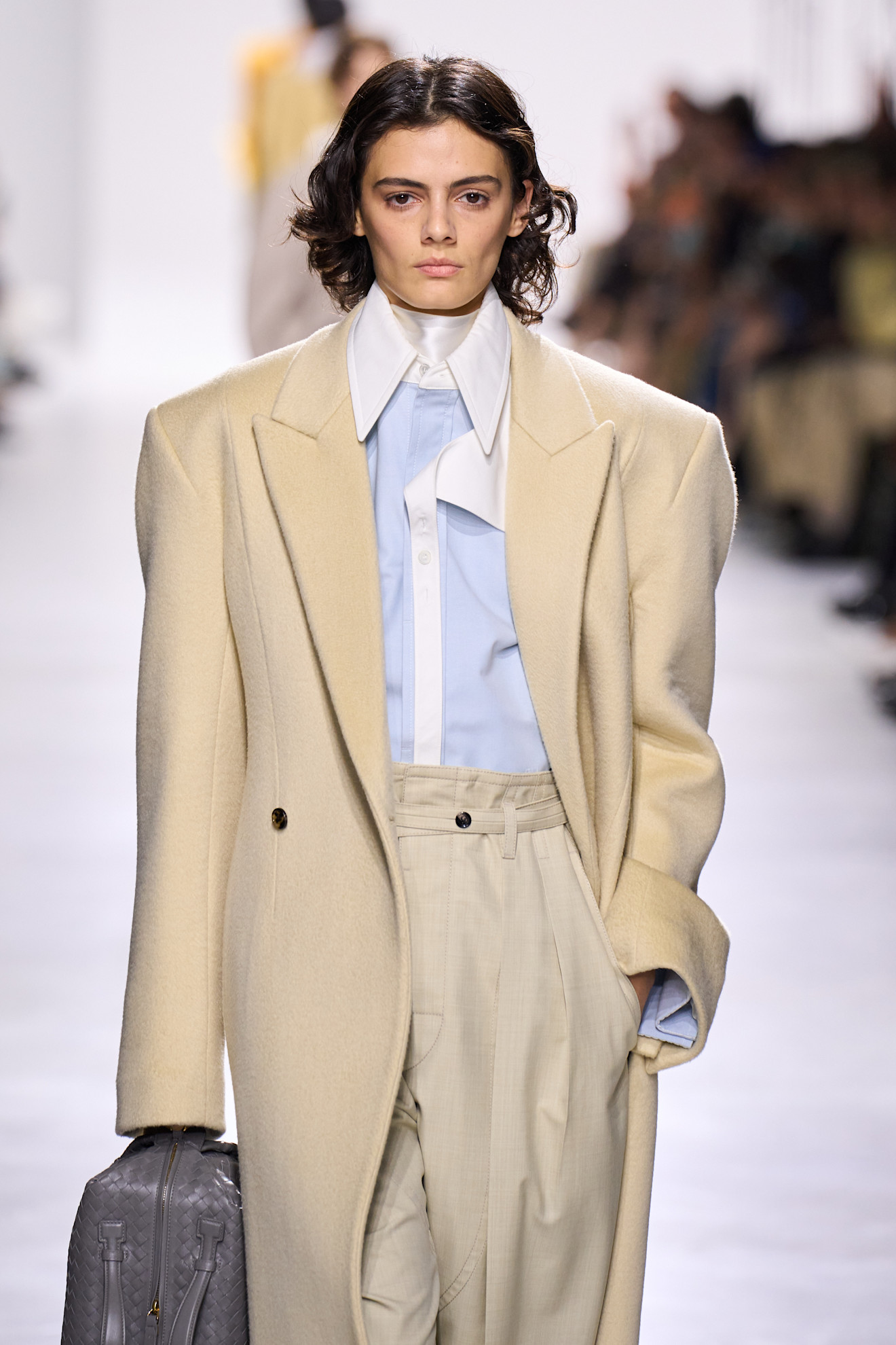 a model at Bottega Veneta Spring 2026 wearing an ivory coat, blue and white button-down, white silk scarf, and tan pants
