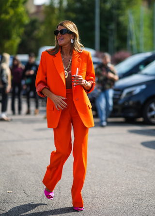 Anna Dello Russo wears black sunglasses, large silver and diamond earrings, a gold large chain with red stones and large pendant necklace, a pale pink lace bra underwear, a gold watch, a neon orange long oversized blazer jacket, matching high waist orange neon flared suit pants, neon pink shiny leather pumps heels shoes with open toe-cap, outside the BOSS fashion show during the Milan Fashion Week - Spring / Summer 2022 on September 23, 2021 in Milan, Italy