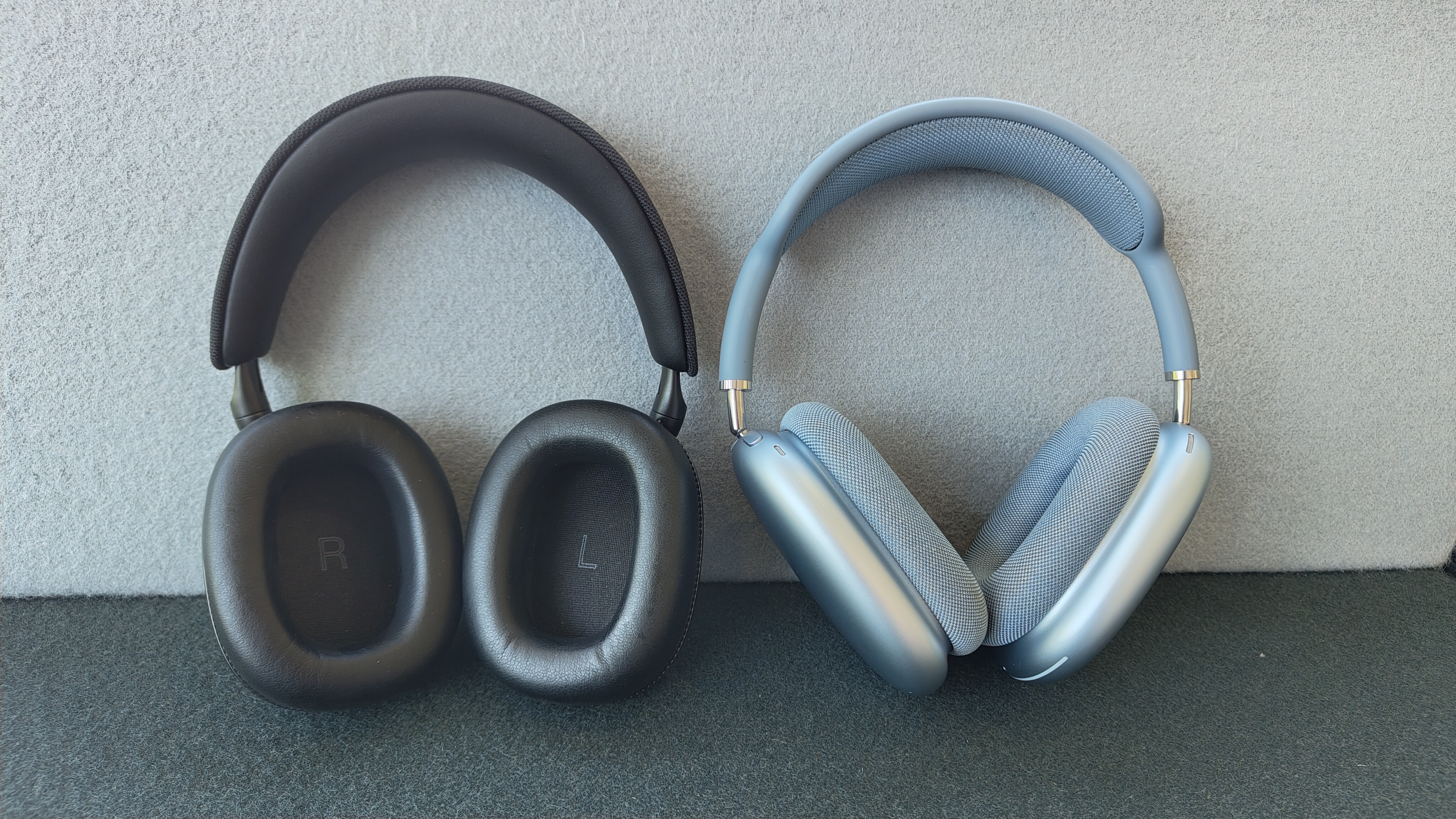 A black pair of B&W Px7 S3 headphones (left) propped up against a wall next to a blue pair of Apple AirPods Max headphones (right). The insides of the Px7 S3's earcups are visible.