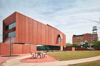 Daytime image of the new contemporary Art Centre, red exterior, grass lawn, circular patio area, paved pathway, table and chairs, surrounding trees, another red brick building in the distance, blue cloudy sky