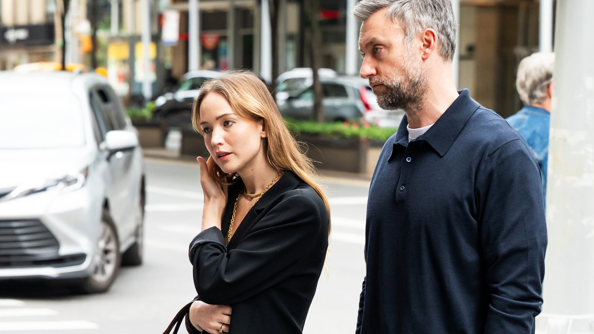 Jennifer Lawrence walking with her husband, wearing a black oversized blazer with a lace-trimmed white slip skirt, strappy nude high-heel sandals, and a croc The Row bag