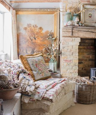 corner of a country cottage living room with armchair covered in vintage fabrics and cushion, antique art on walls and brick fireplace surround