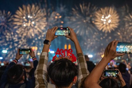 A crowd capturing New Year’s fireworks with their smartphones, with golden fireworks illuminating the night sky.