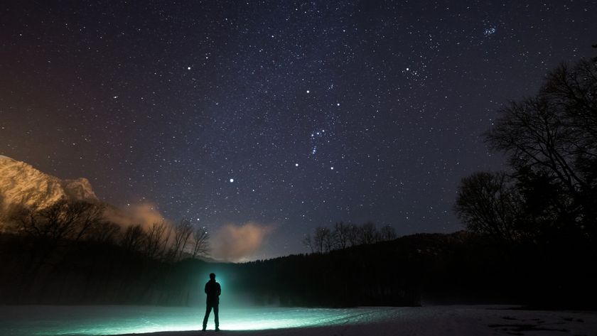 A silhouette of a person stands in front of a green flashlight beam on the edge of a lake with trees and hills in the distance all underneath a dark blue starry sky.