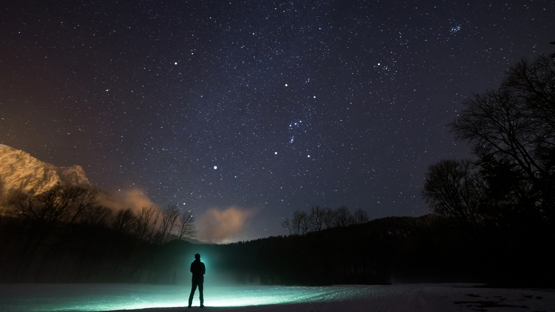 A silhouette of a person stands in front of a green flashlight beam on the edge of a lake with trees and hills in the distance all underneath a dark blue starry sky.