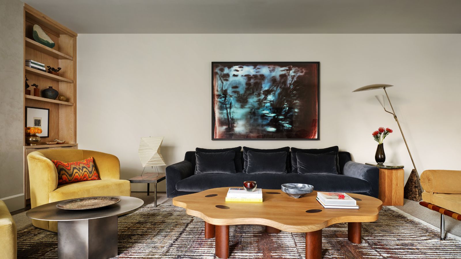 Living room with large rug, curved coffee table, velvet blue sofa, yellow velvet armchair, tall floor lamp and wood bookcase