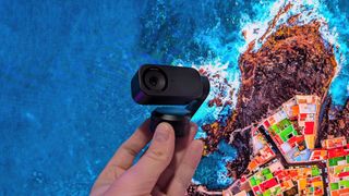 The Insta360 Link 2 webcam held in the hand with a vibrant image of a coastal city in the background.