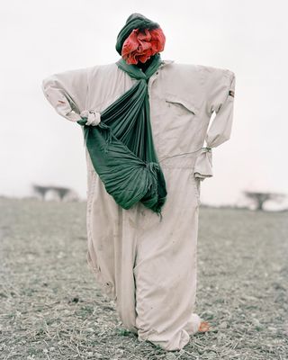 A "scarecrow" dressed in a work coverall in gray, with a green fabric around its neck.