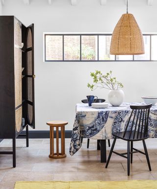 white painted dining room with paneled window, stone flooring, rectangular dining table with blue and white table cloth, low hanging woven pendant, wooden black dining chair and wooden stool, black wood and rattan unit beside table, yellow rug