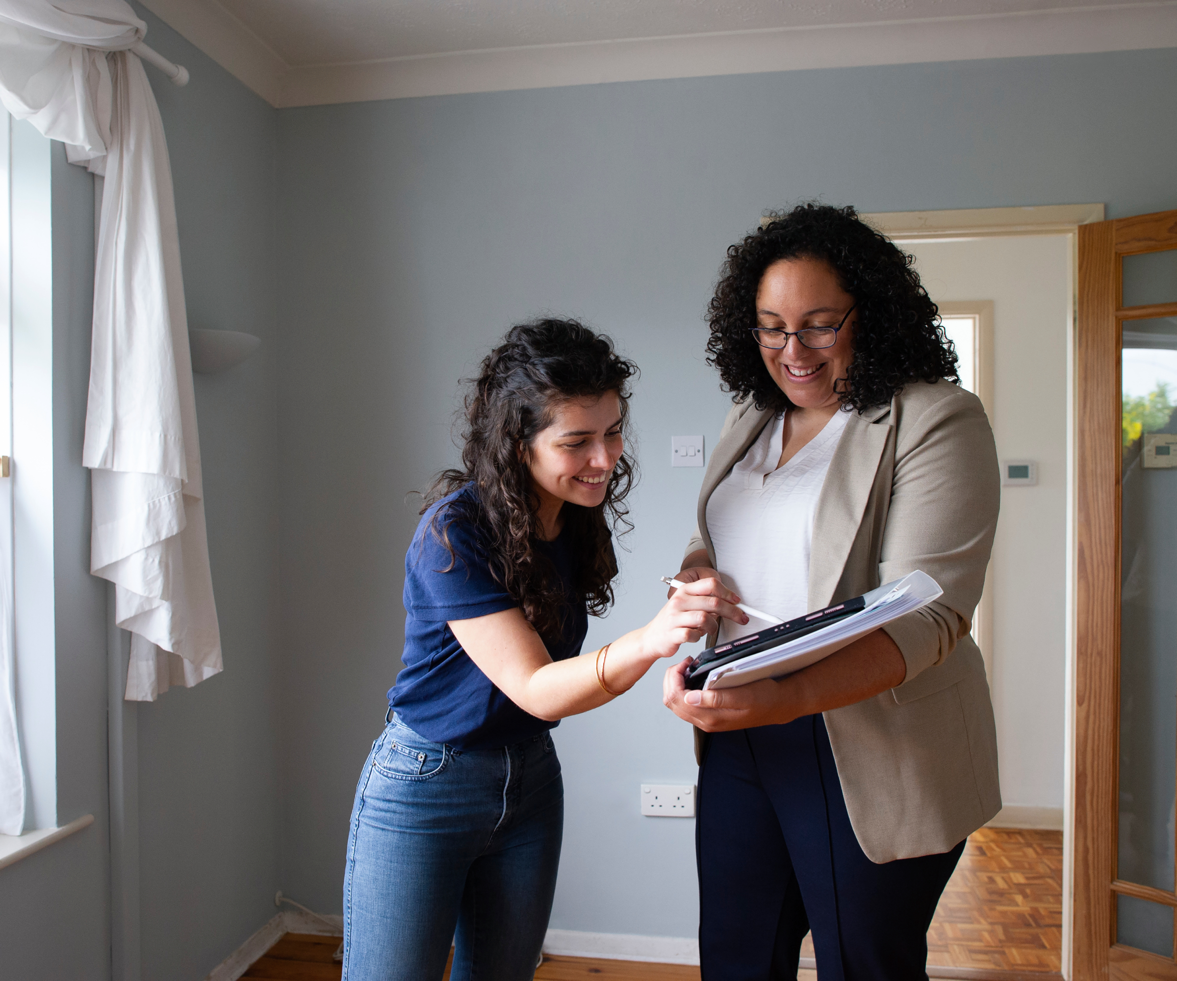 female signing document being held by another female, both stood in empty room of house