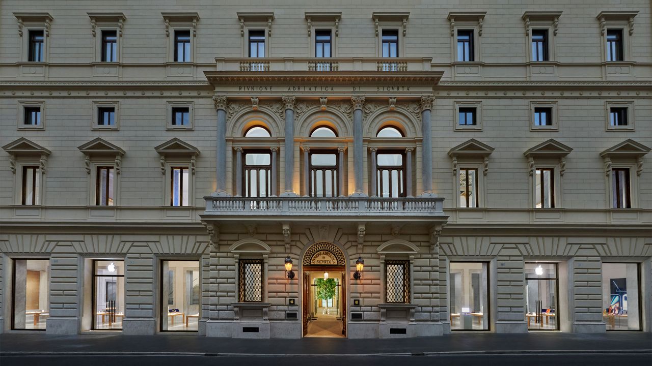 The front of a three storey building with large windows on the street level, a balcony in the middle of the building on the second level which is surrounded by rows of windows on the other levels.