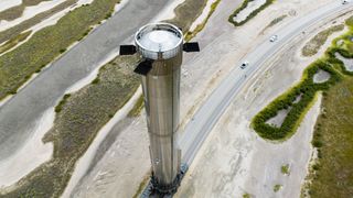a tall silver rocket booster is rolled down a road surrounded by wetlands and sand dunes.