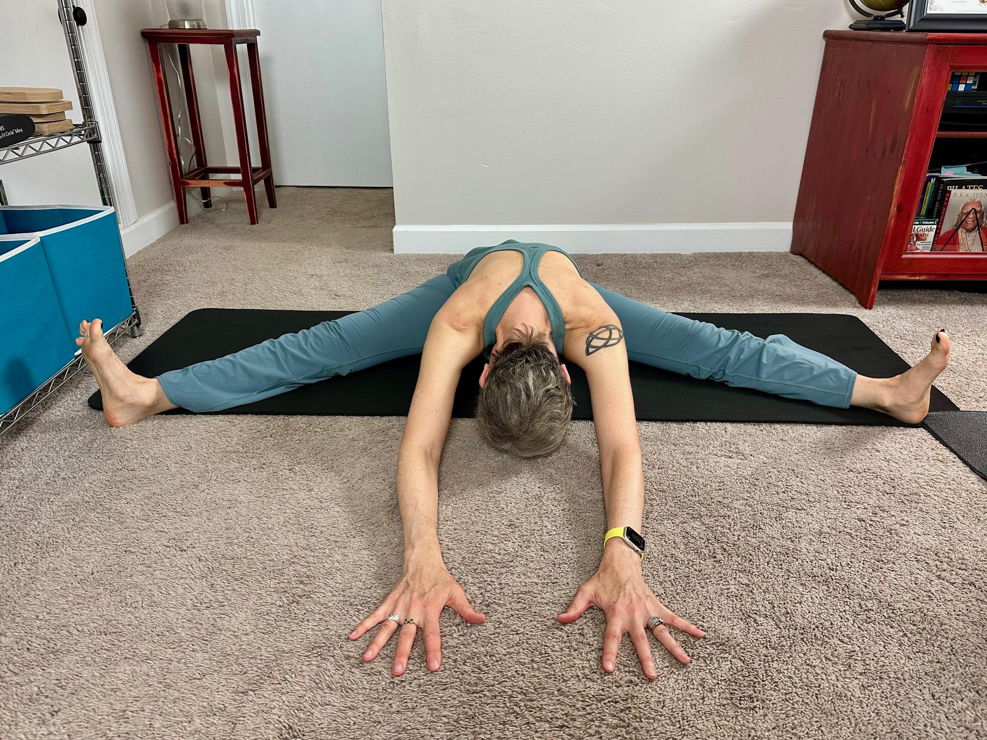 woman seated on the floor on a black yoga mat, legs wide folding forward
