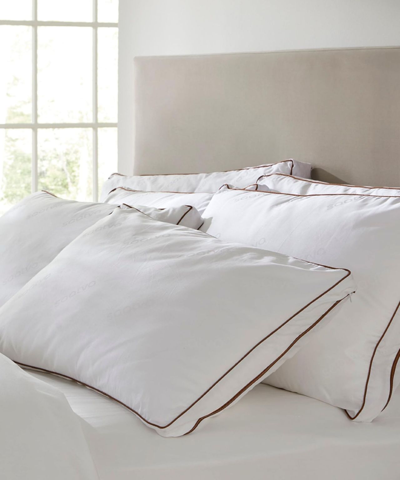 Saatva Latex pillows stacked up in a white bedroom with a tall beige headboard and windows seen beyond