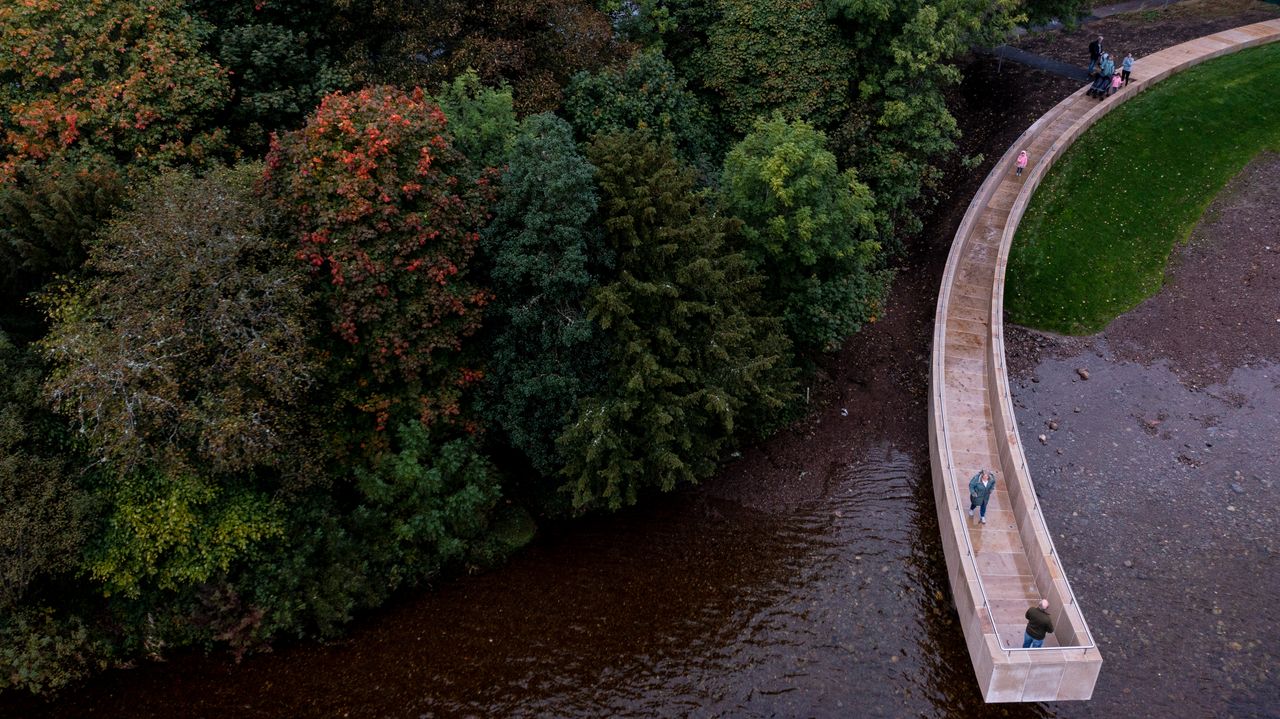 Daytime image of The gathering place installation in Scotland, stone curved pathway suspended over the water, people walking on the path, forest area to the left, grass verge to the left