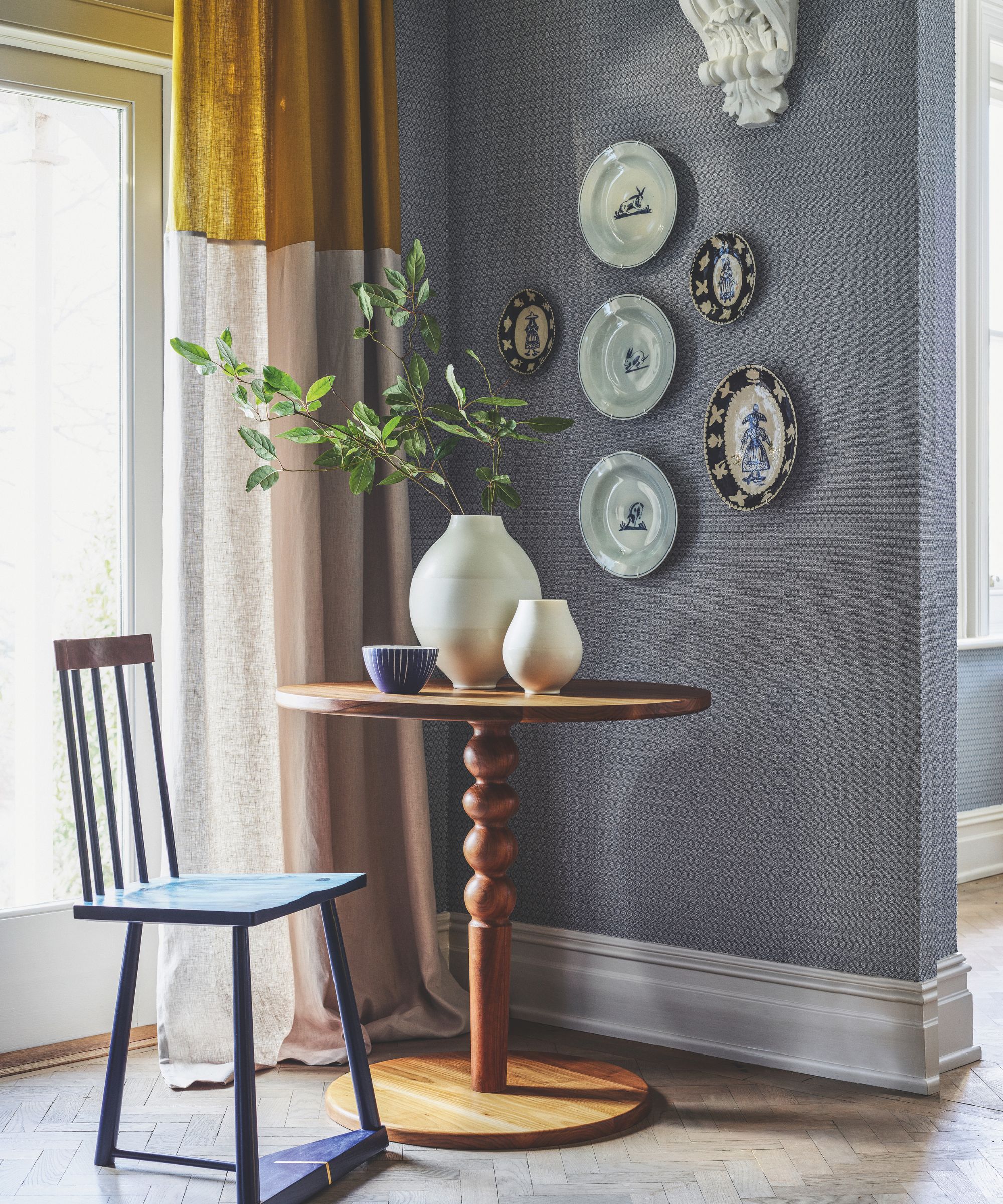 A round wooden table with a chair, vases, and decorative plates on a wallpapered wall. Behind the chair is a bright window with a cream and ochre curtain.
